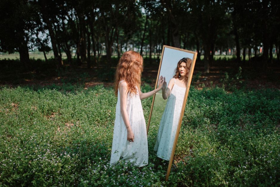 Young woman in a white dress gazing at her reflection in a mirror amid a lush green park setting.
