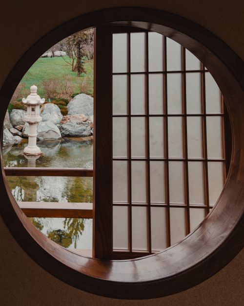 Free A tranquil garden scene viewed through a traditional Japanese circular window, with a stone lantern and pond. Stock Photo