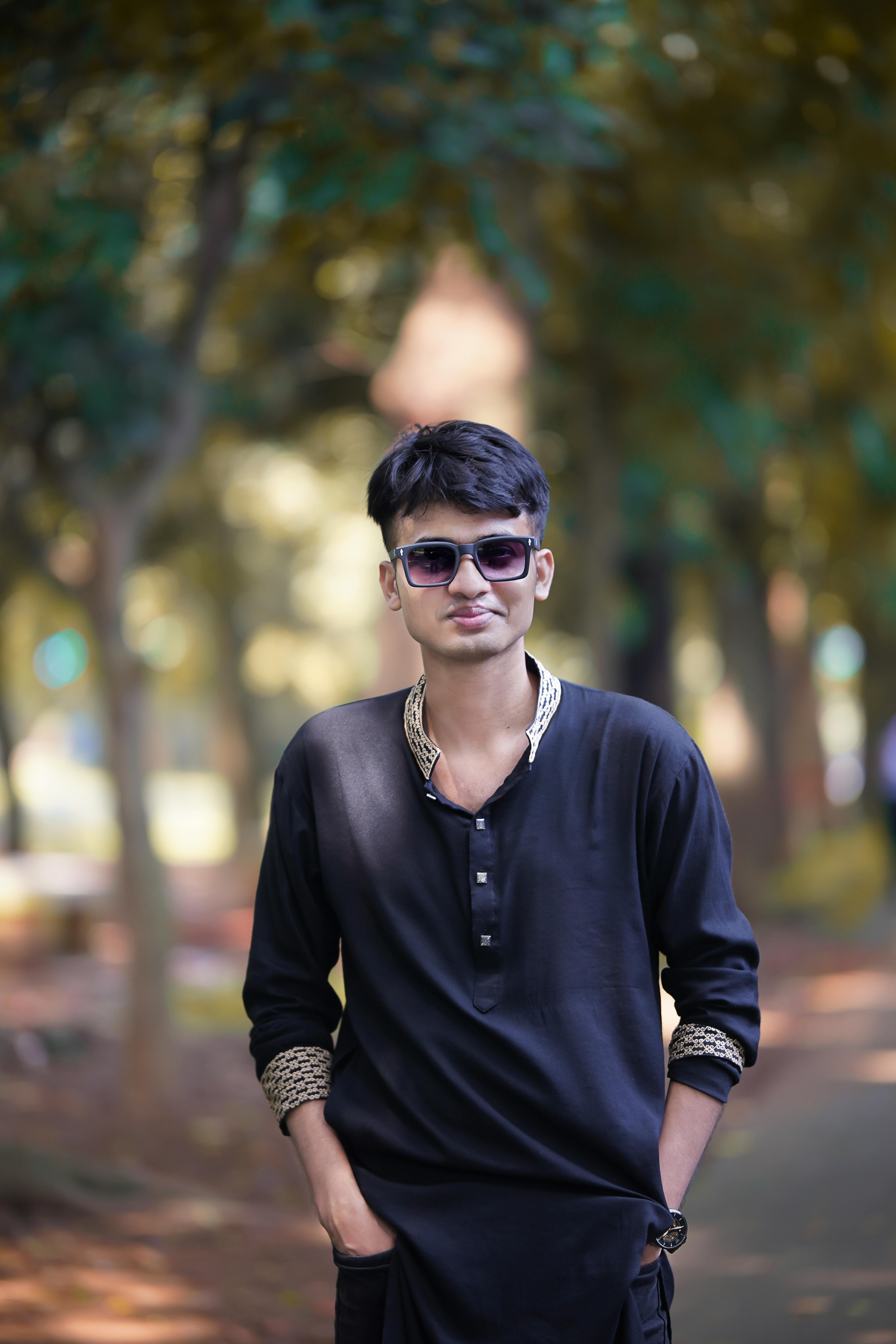 Stylish young man wearing sunglasses and black attire, standing confidently on a forest path.