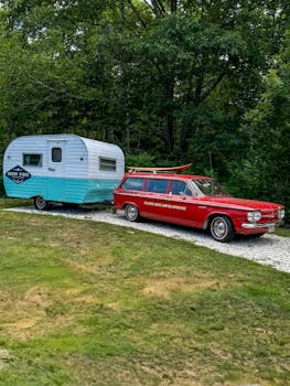 Classic red car with a vintage camper parked on a gravel path in a lush green setting.