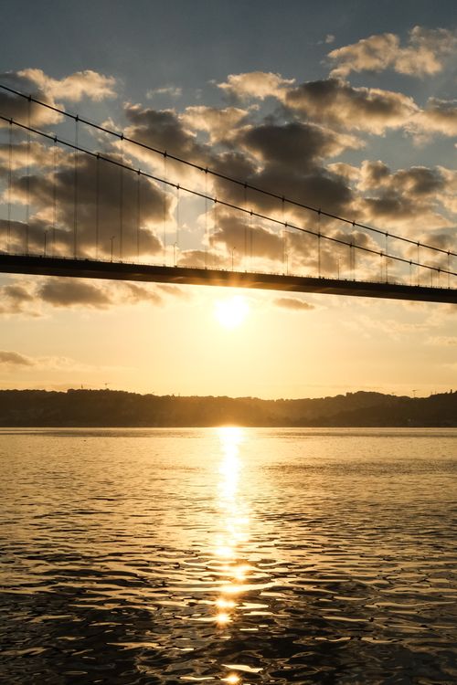Free Sunset view of a suspension bridge silhouette reflecting on calm water with clouds. Stock Photo