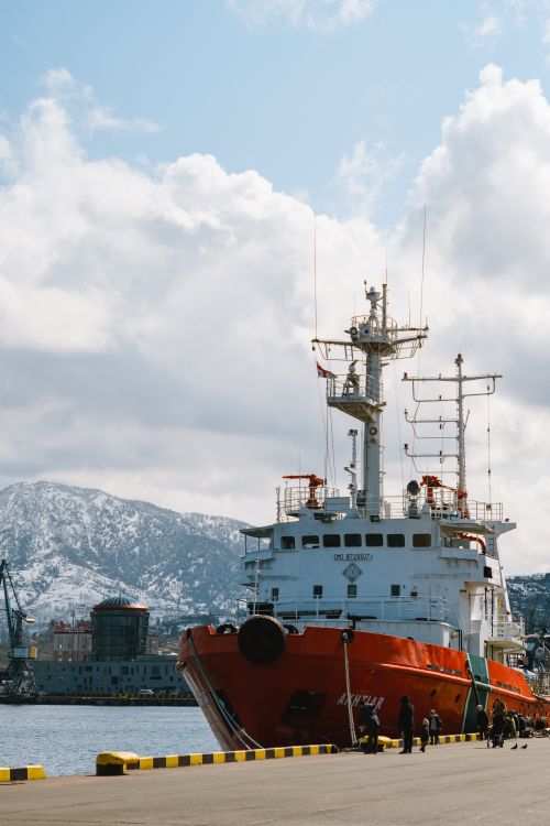 Free A vibrant red ship docked at a harbor with scenic mountain views and clear skies, capturing a moment of serene industrial activity. Stock Photo