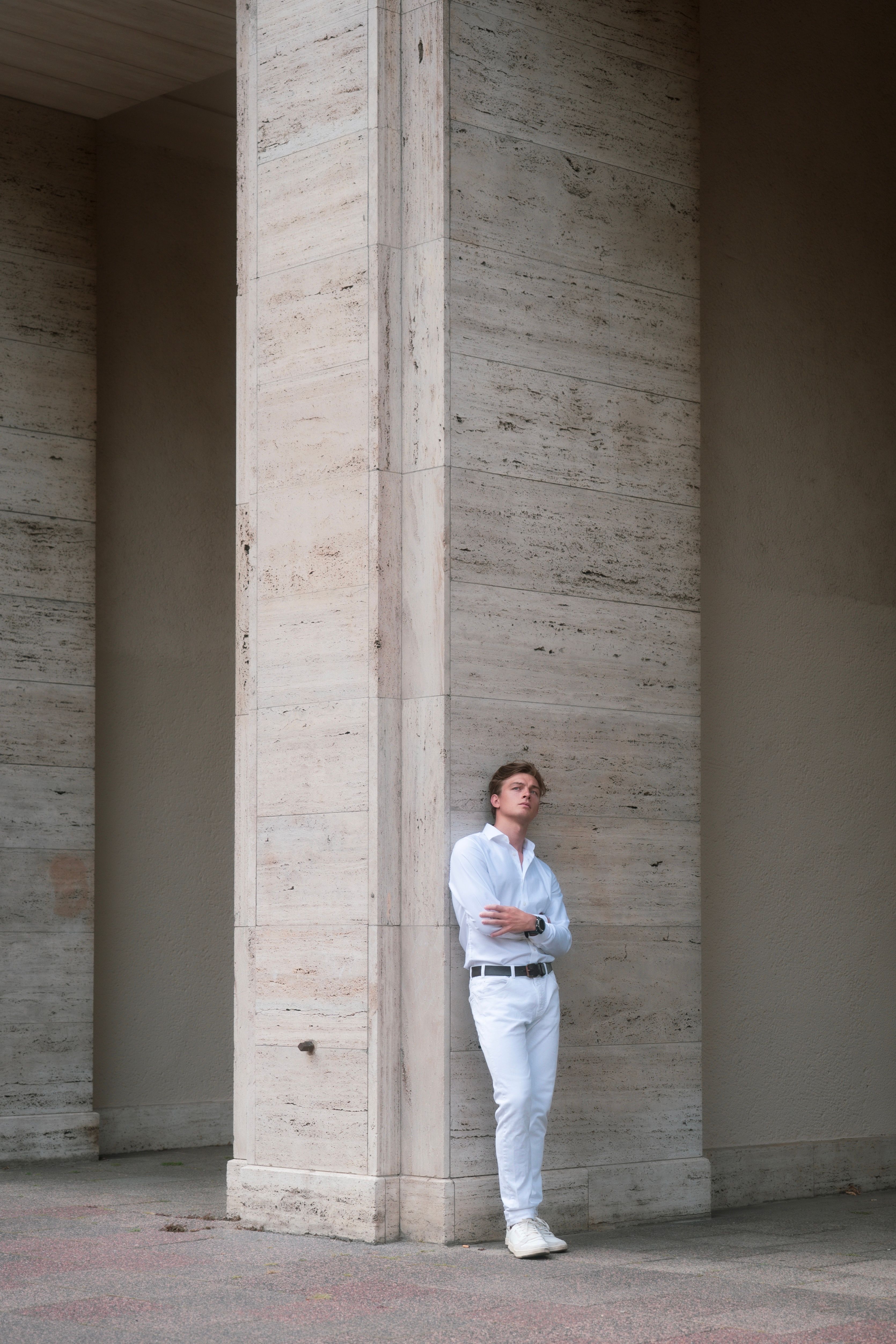 A fashionable man in white leans against a stone column in Berlin, displaying modern architecture.