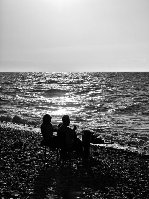 Free A black and white image capturing a couple silhouetted against the sunset over the sea, providing a serene atmosphere. Stock Photo