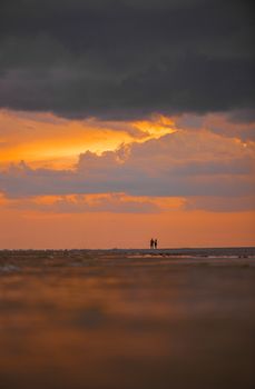 Silhouette of two people walking along the beach during a vibrant sunset with dramatic clouds.