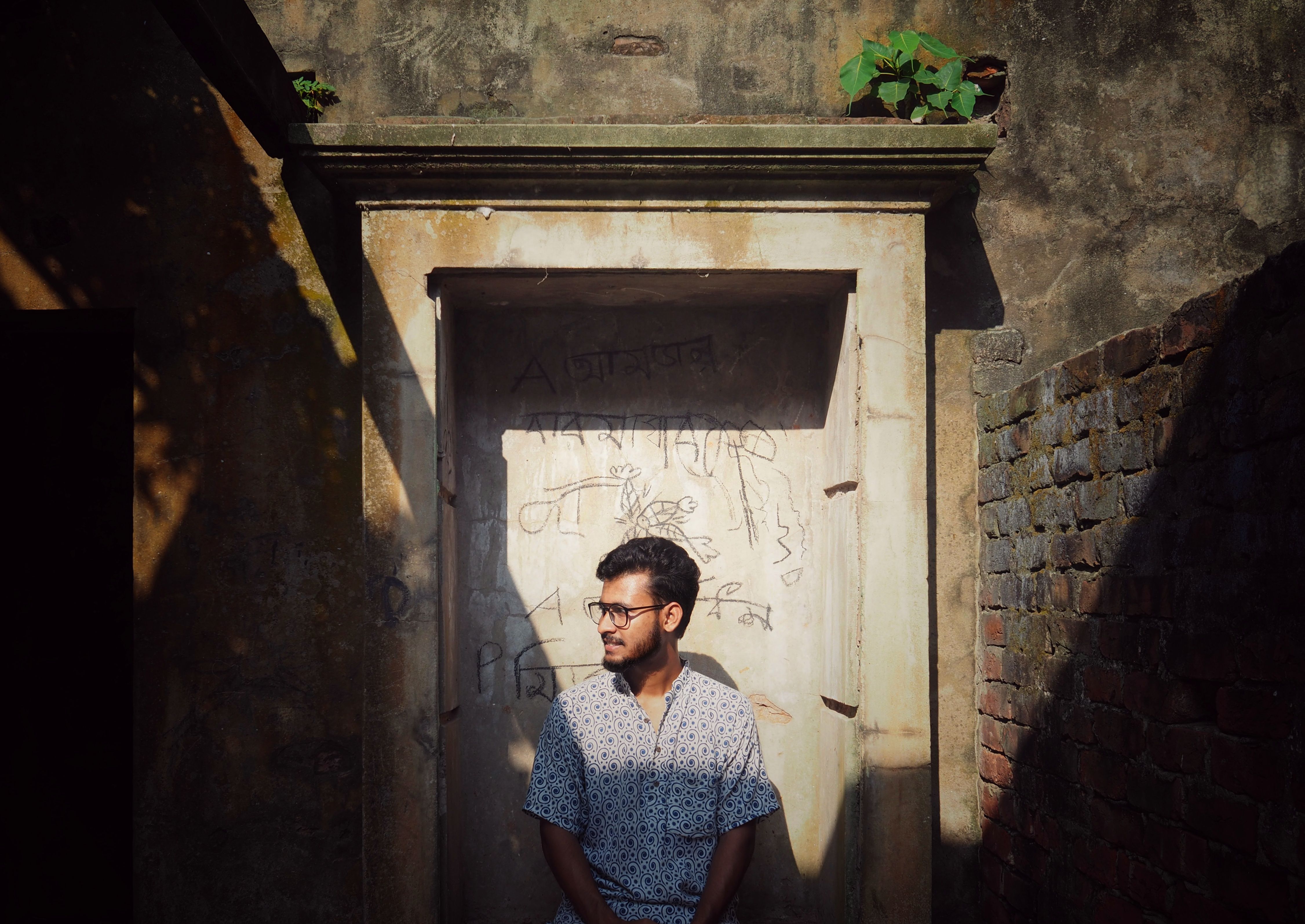 A young man with eyeglasses stands in a sunlit doorway surrounded by brick walls.