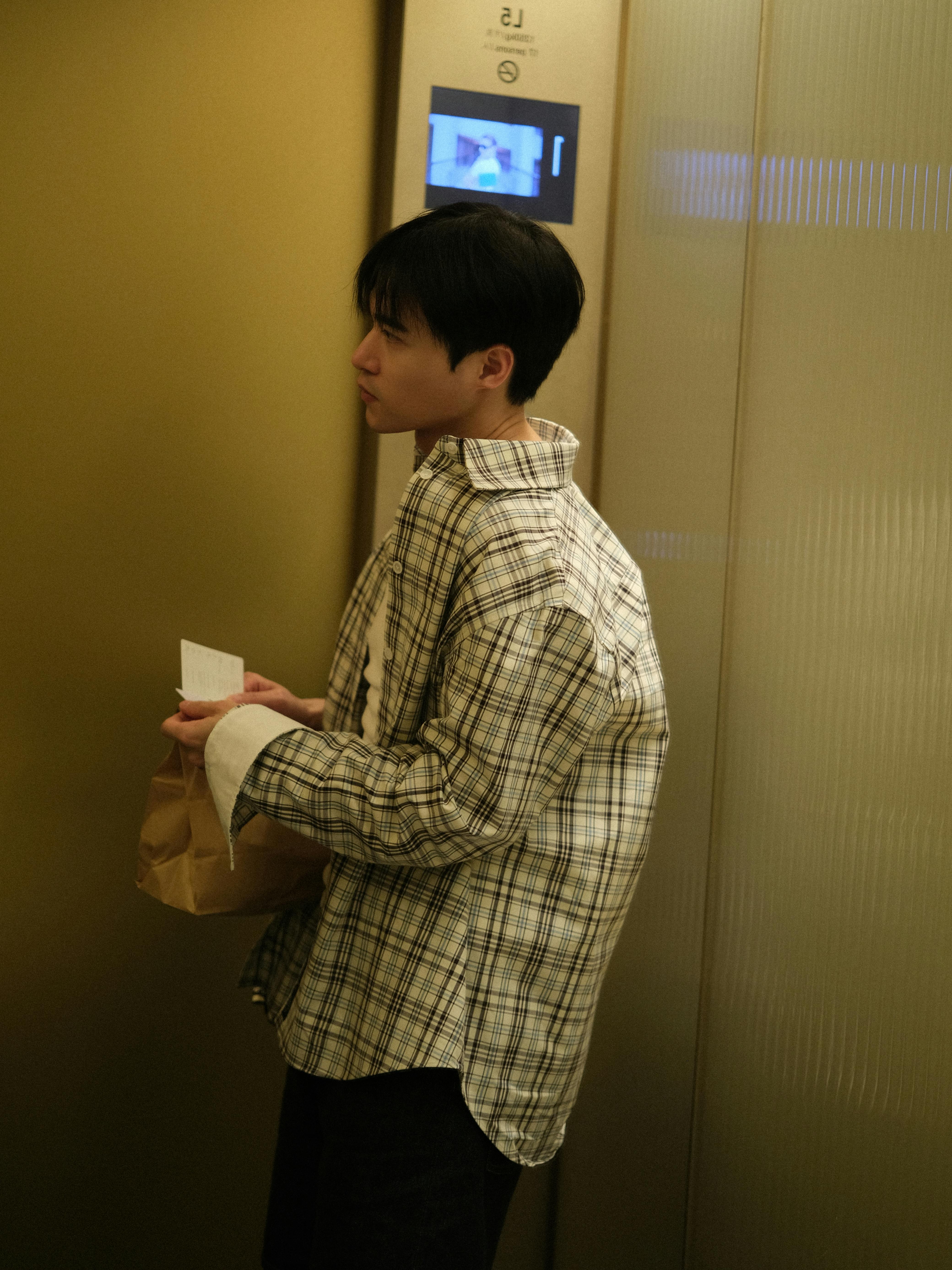 Young man in a plaid shirt waiting in an elevator lobby holding a paper bag.