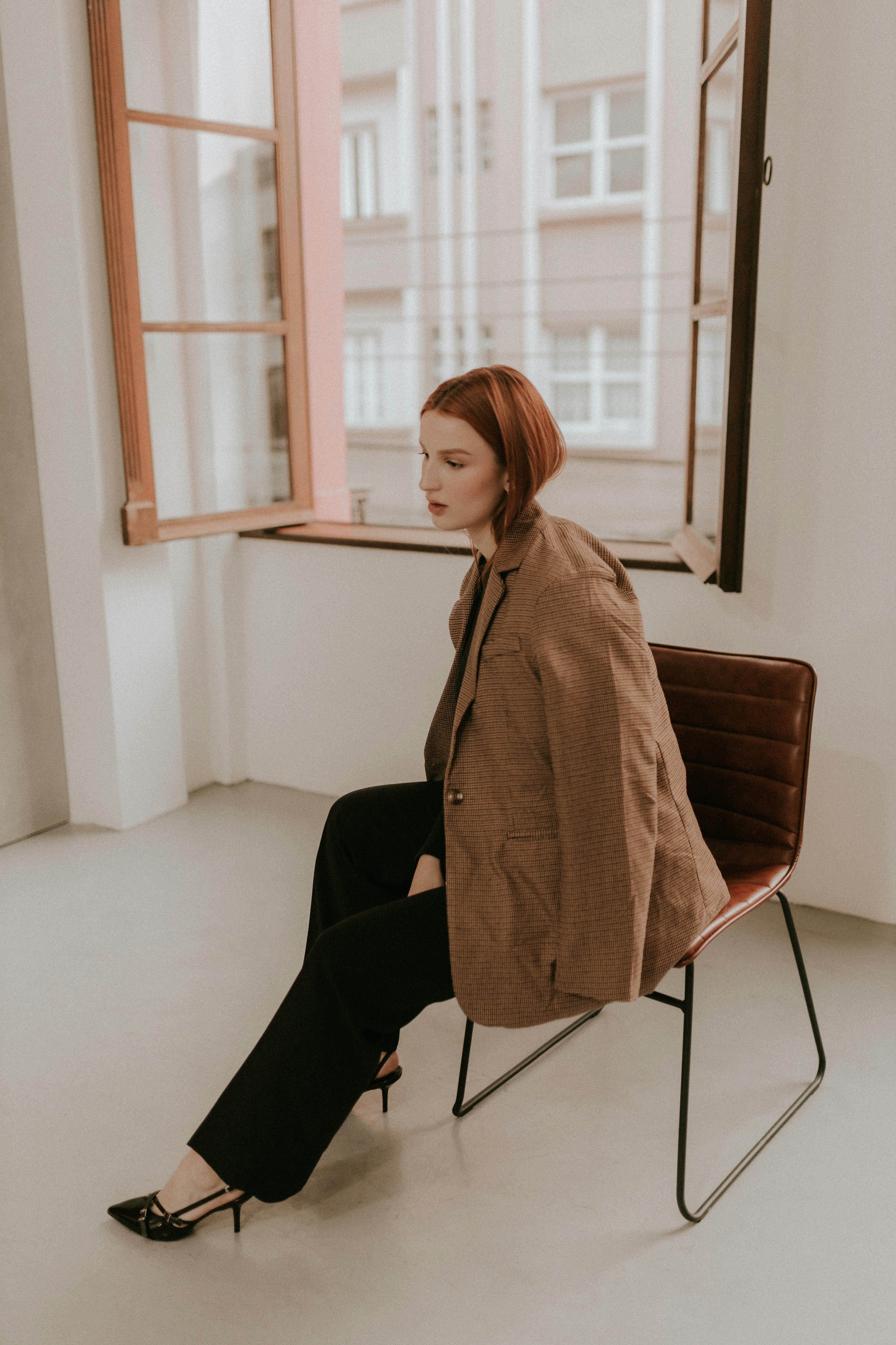 Fashionable woman in blazer sitting indoors near open window in modern space.