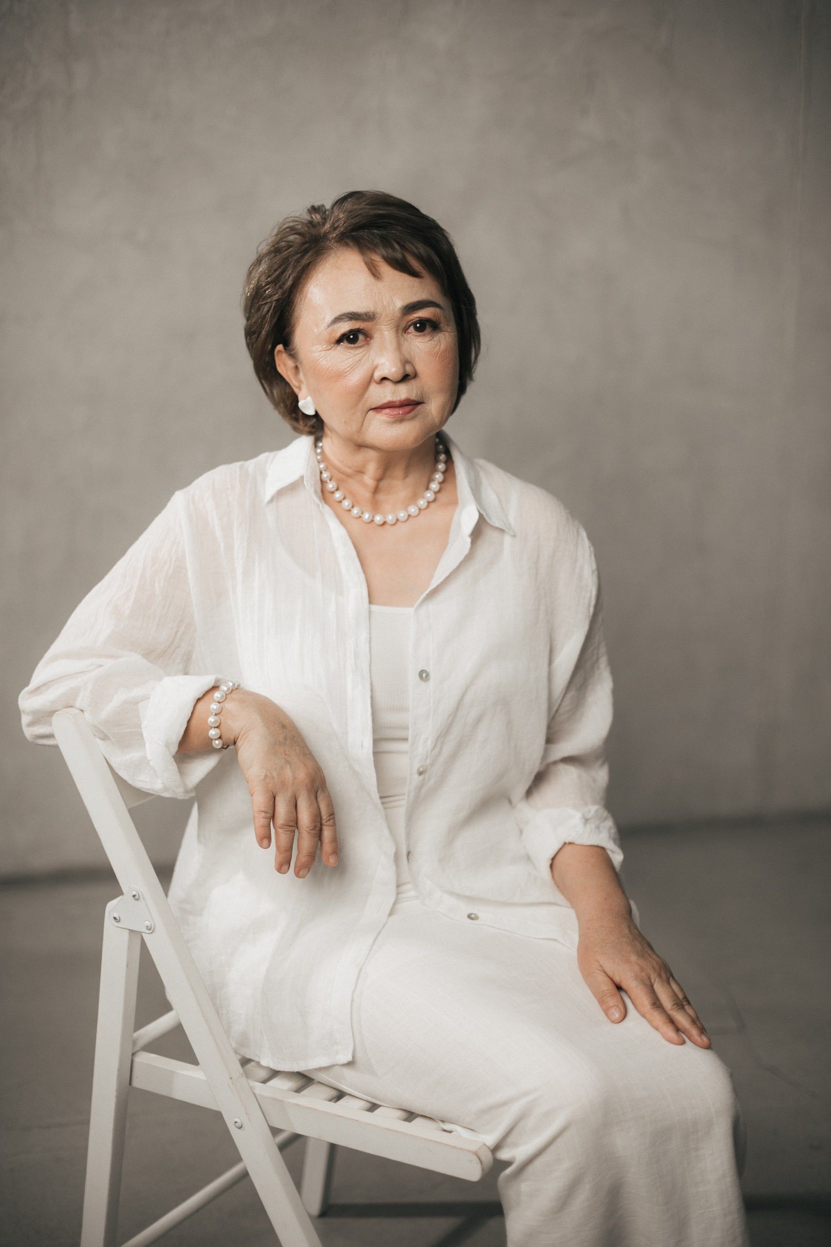 Graceful senior woman in a white outfit sitting on a chair, in a studio setting.