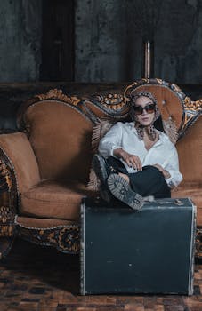 Chic woman in eyewear and scarf sitting on ornate vintage sofa with suitcase.
