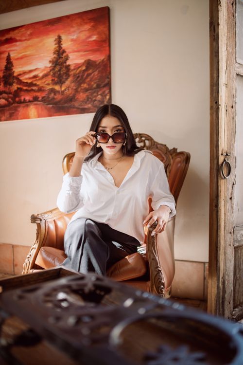 Free Elegant woman in a white shirt and sunglasses sitting on a vintage chair indoors. Stock Photo