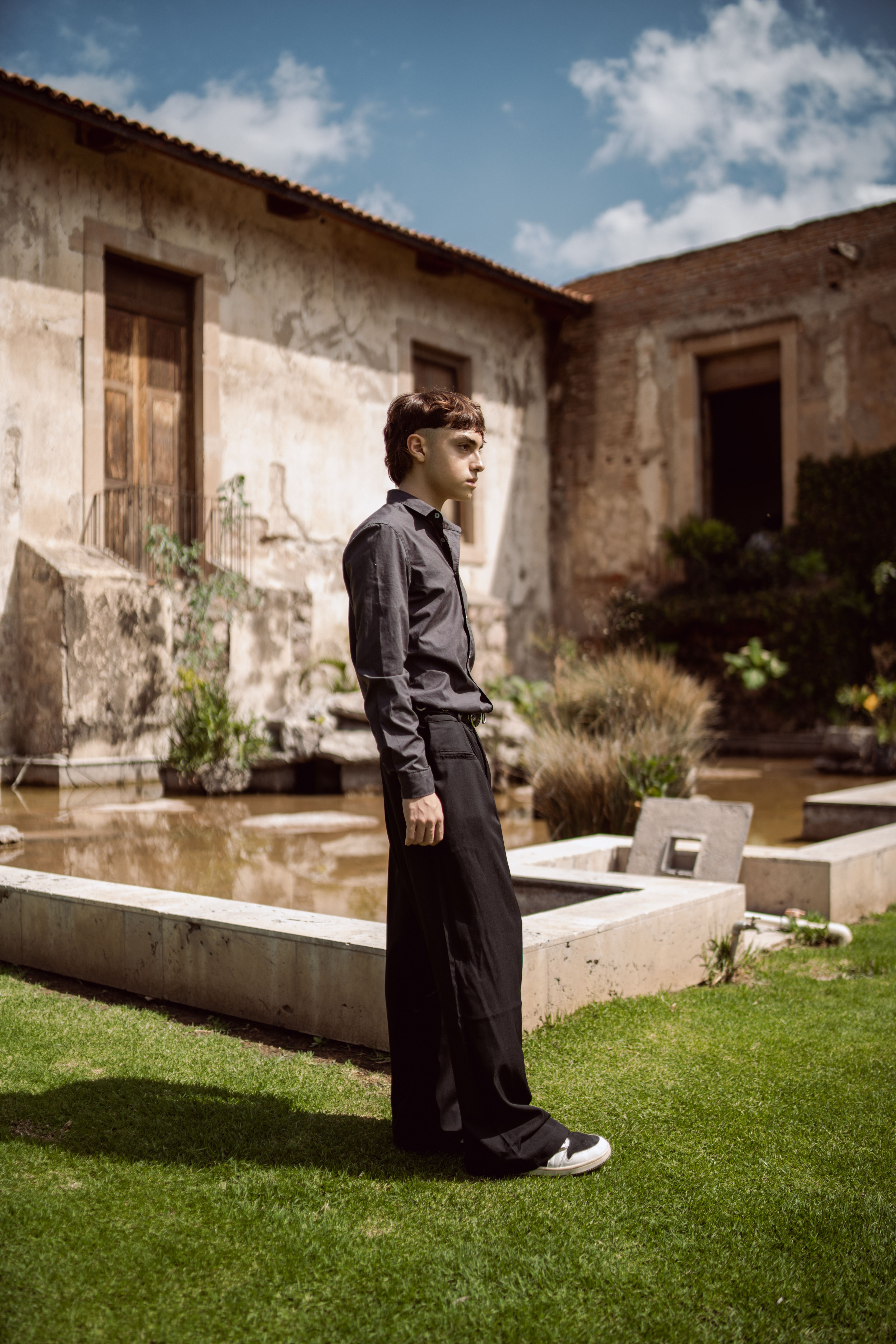 Fashion model in black attire stands in a rustic courtyard, showcasing timeless style.