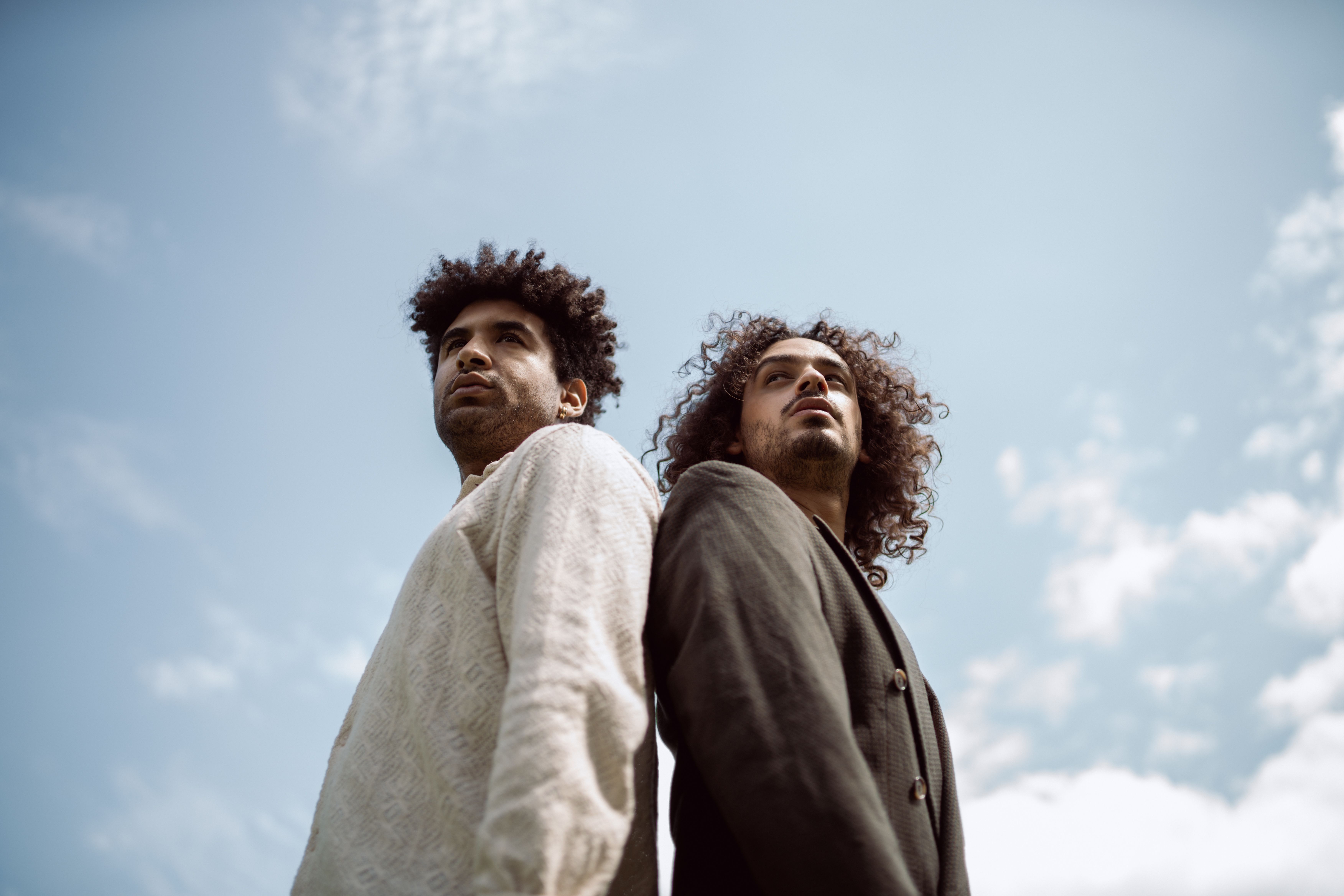 Two stylish men with curly hair posing outdoors against a bright blue sky.