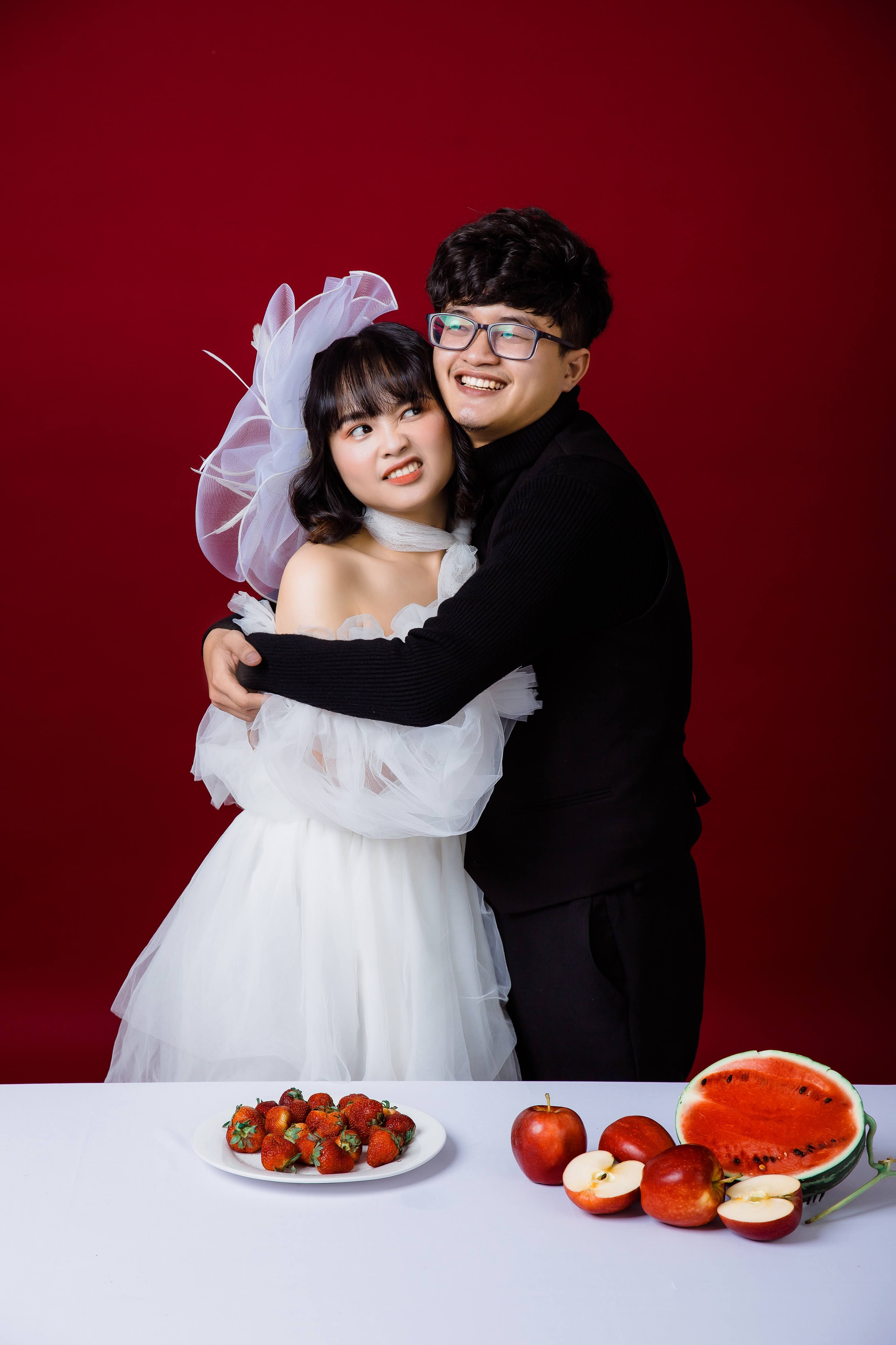 Free Joyful couple in wedding attire embracing with fruit on table against red background. Stock Photo