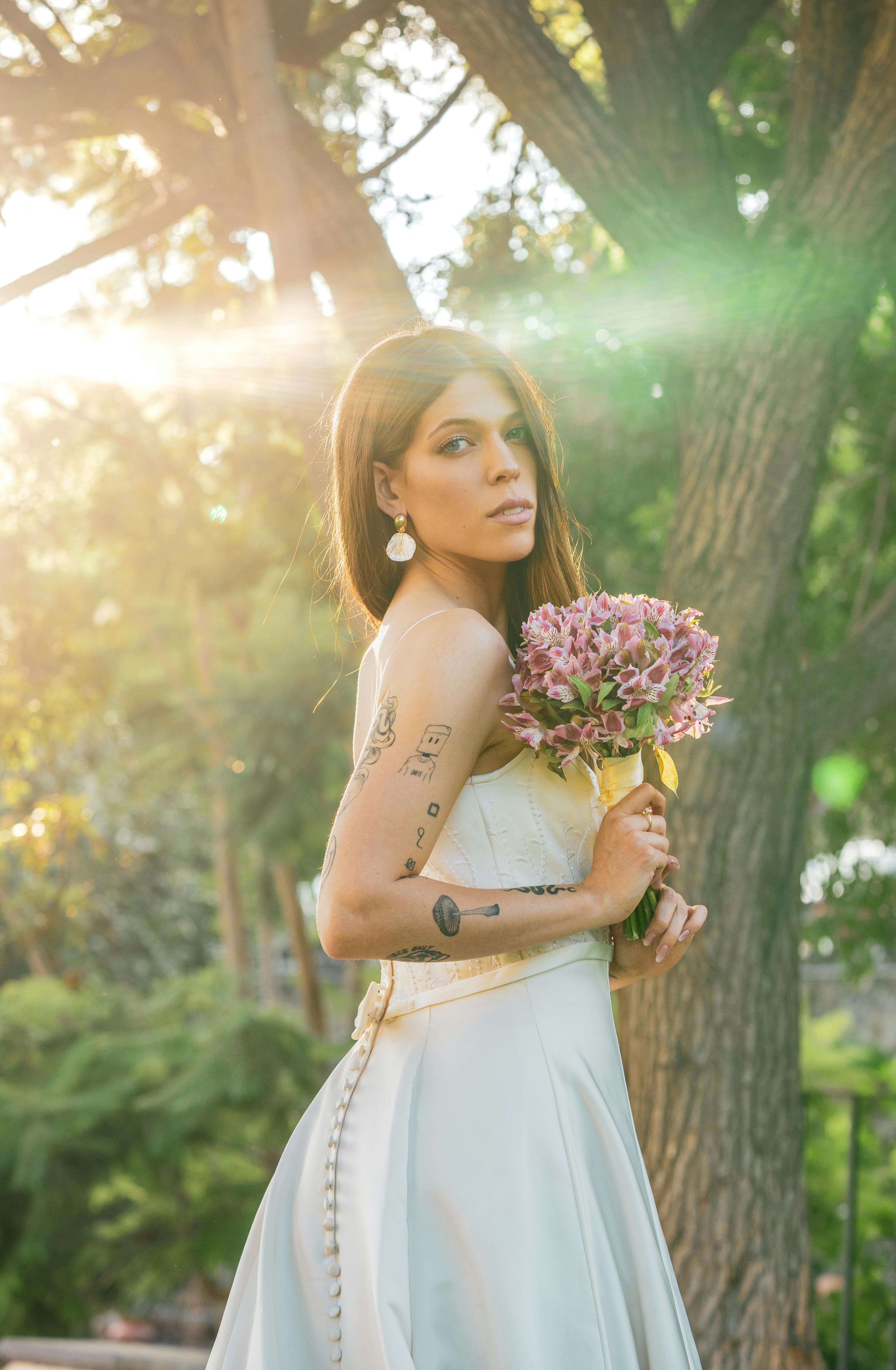 A beautiful bride in a white gown holds a bouquet under warm sunlight in a park setting.