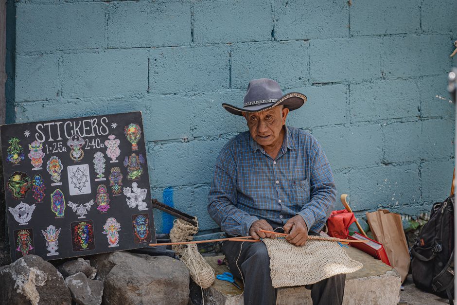 An elderly man in Panajachel, Guatemala, creates woven products surrounded by colorful stickers at a street market.