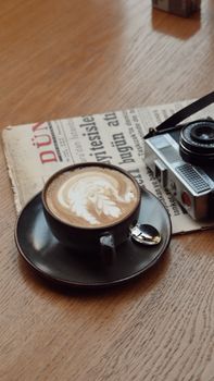 High-angle view of a cappuccino with intricate latte art beside a vintage camera and newspaper.