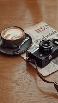 A vintage camera, cappuccino, and newspaper on a wooden table evoke a nostalgic vibe.