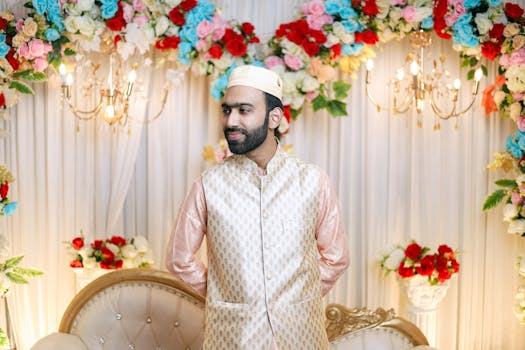 Groom in embroidered attire posing at a vibrant, floral wedding setting indoors.