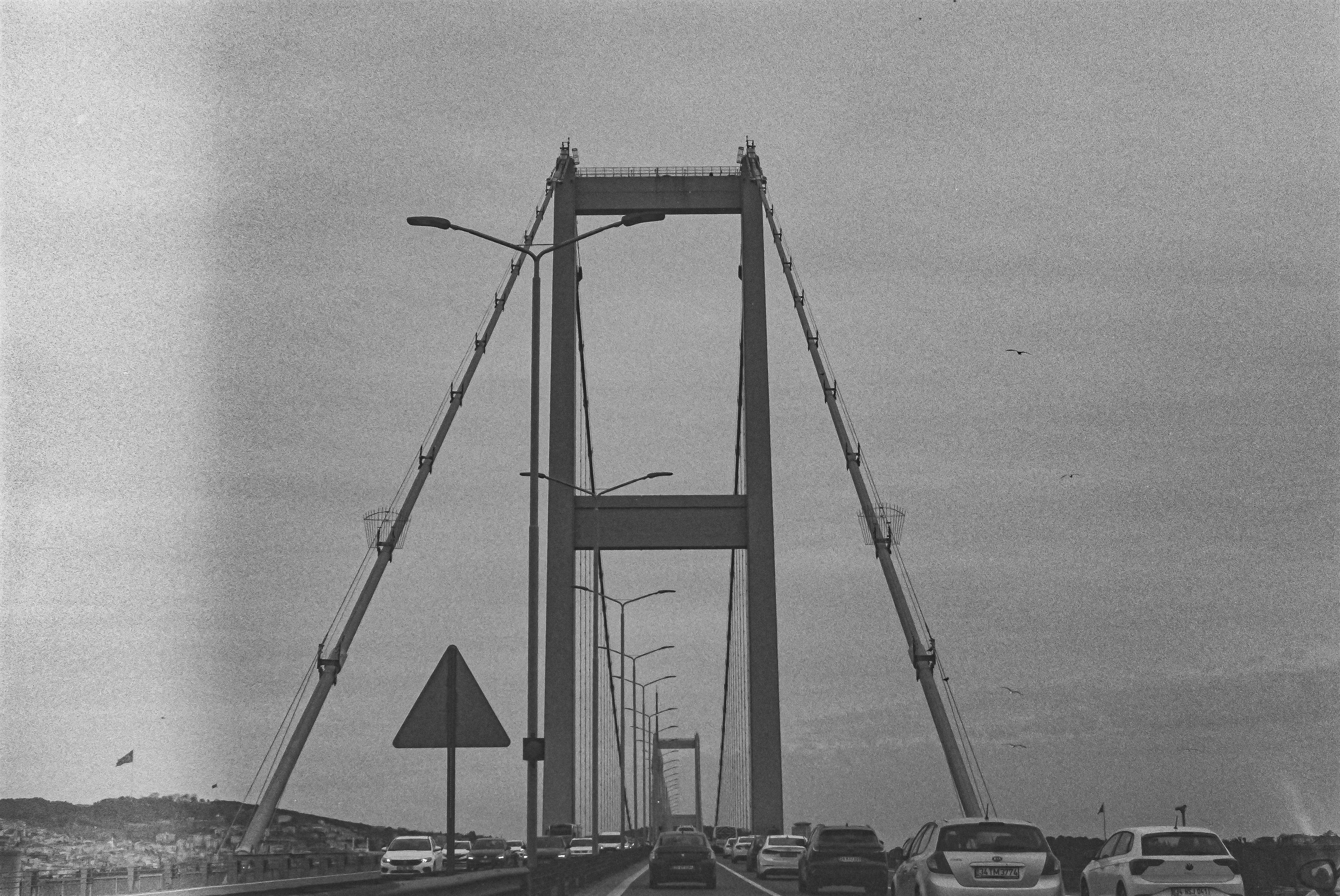 Monochrome image depicting a suspension bridge in an industrial area with vehicles.