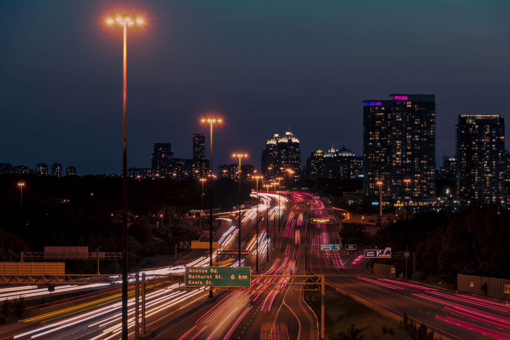 Light trails along a Greater Toronto Area highway at dusk with the city skyline beyond.