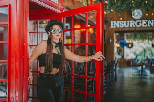 Free Trendy woman in black outfit standing confidently by a red phone booth, enhancing urban aesthetics. Stock Photo