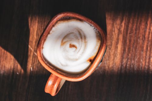 Free Overhead view of creamy cappuccino in a rustic ceramic mug on a wooden table, sunlight highlighting foam. Stock Photo