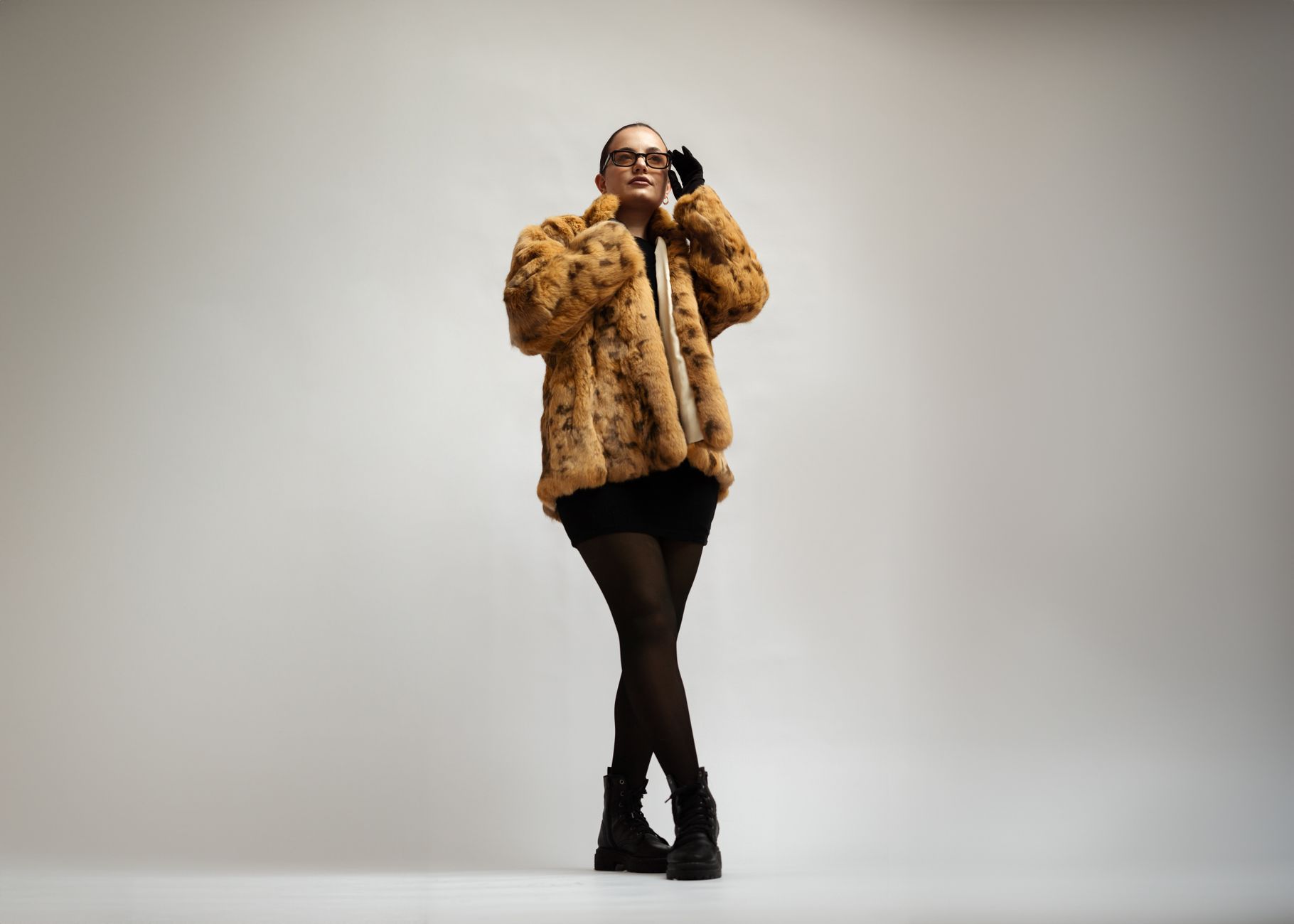 Stylish young woman in fur coat and glasses posing in a studio setting.