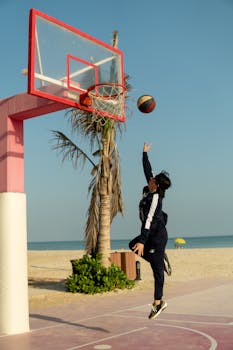 A young man jumps to shoot a basketball on a sandy beach court at sunrise.