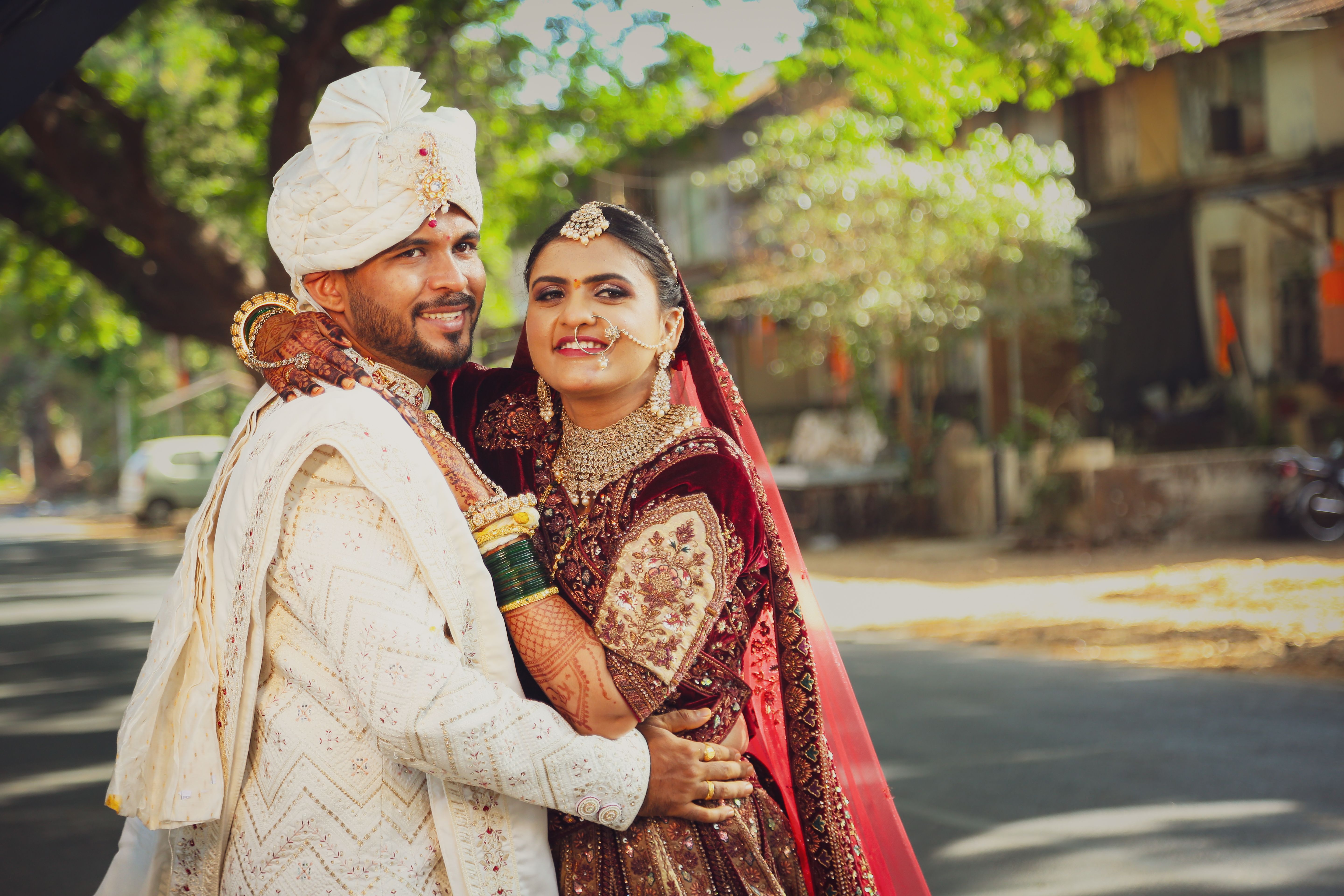 Joyful couple in traditional wedding attire embracing outdoors, surrounded by street scene and trees.
