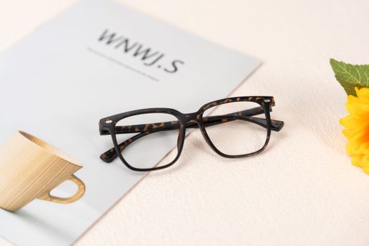 Tortoise-shell eyeglasses on a minimalist desk with magazine, bright cup, and vibrant flower.