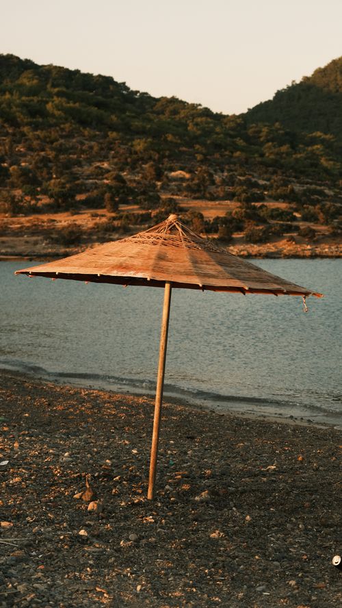 Free A serene beach scene featuring a solitary straw umbrella against a sunset backdrop by the ocean. Stock Photo