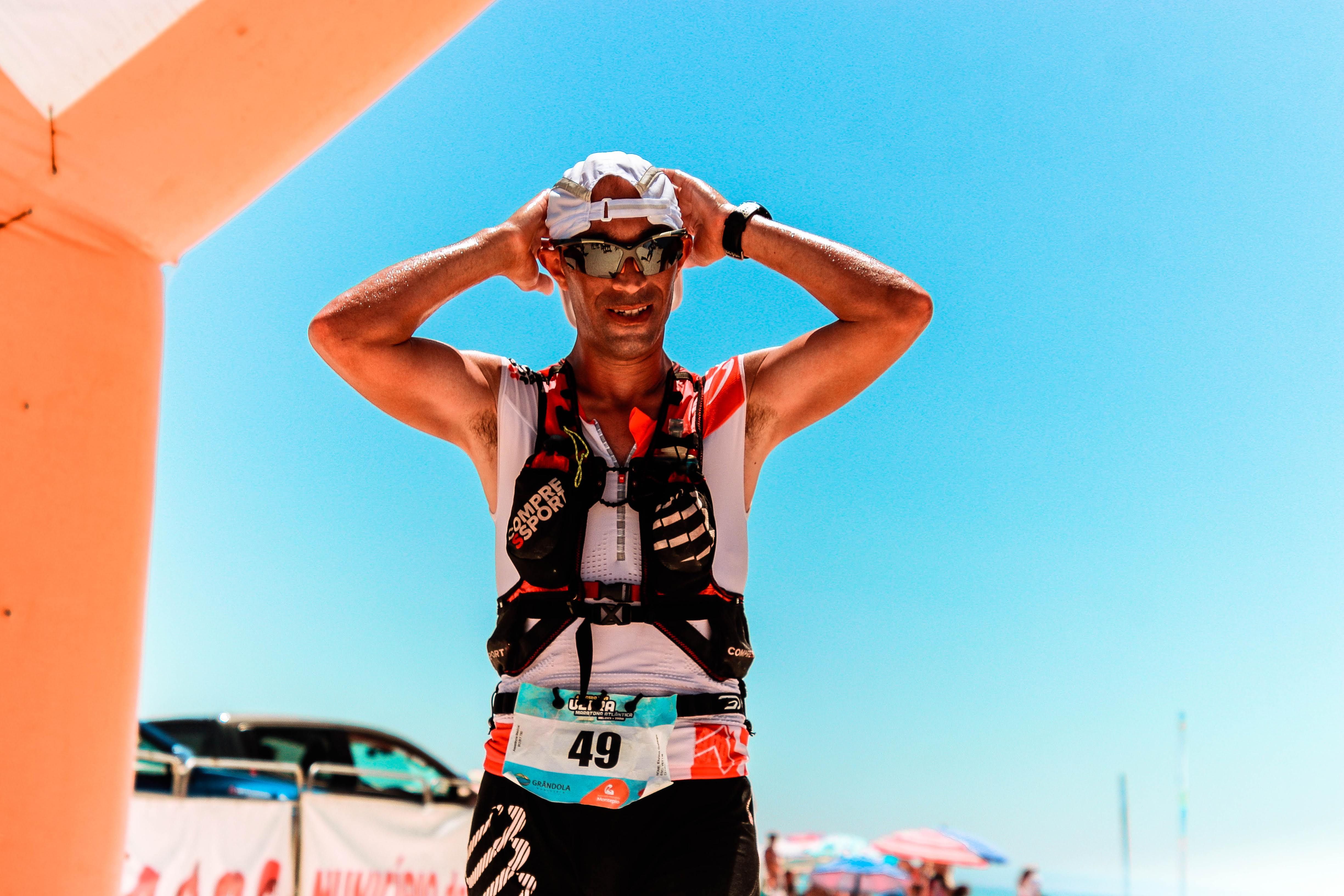 Smiling male athlete wearing sunglasses and cap at beach marathon finish line under blue sky.