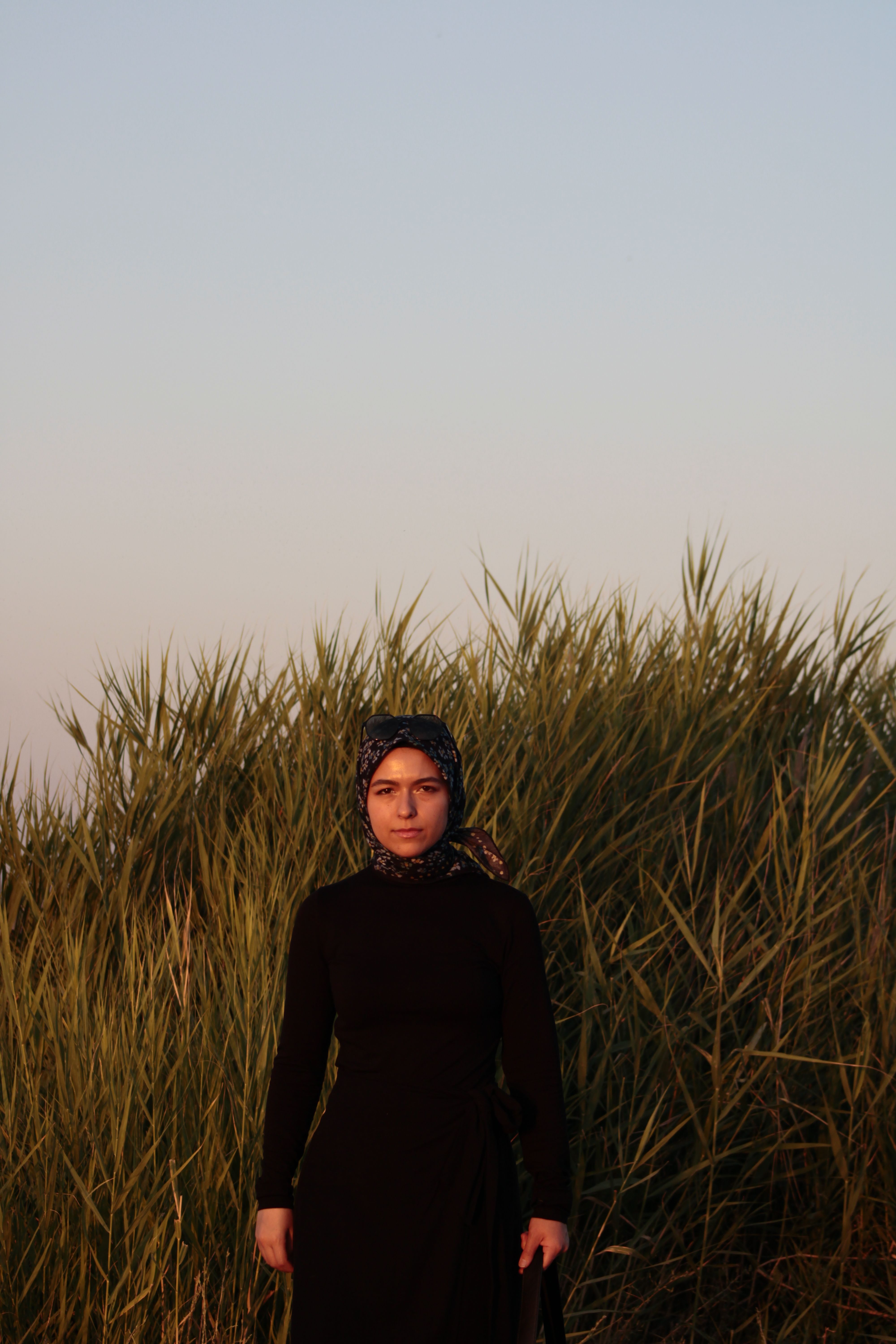 A woman in a black dress stands in front of tall grass during sunset, exuding a serene and peaceful vibe.