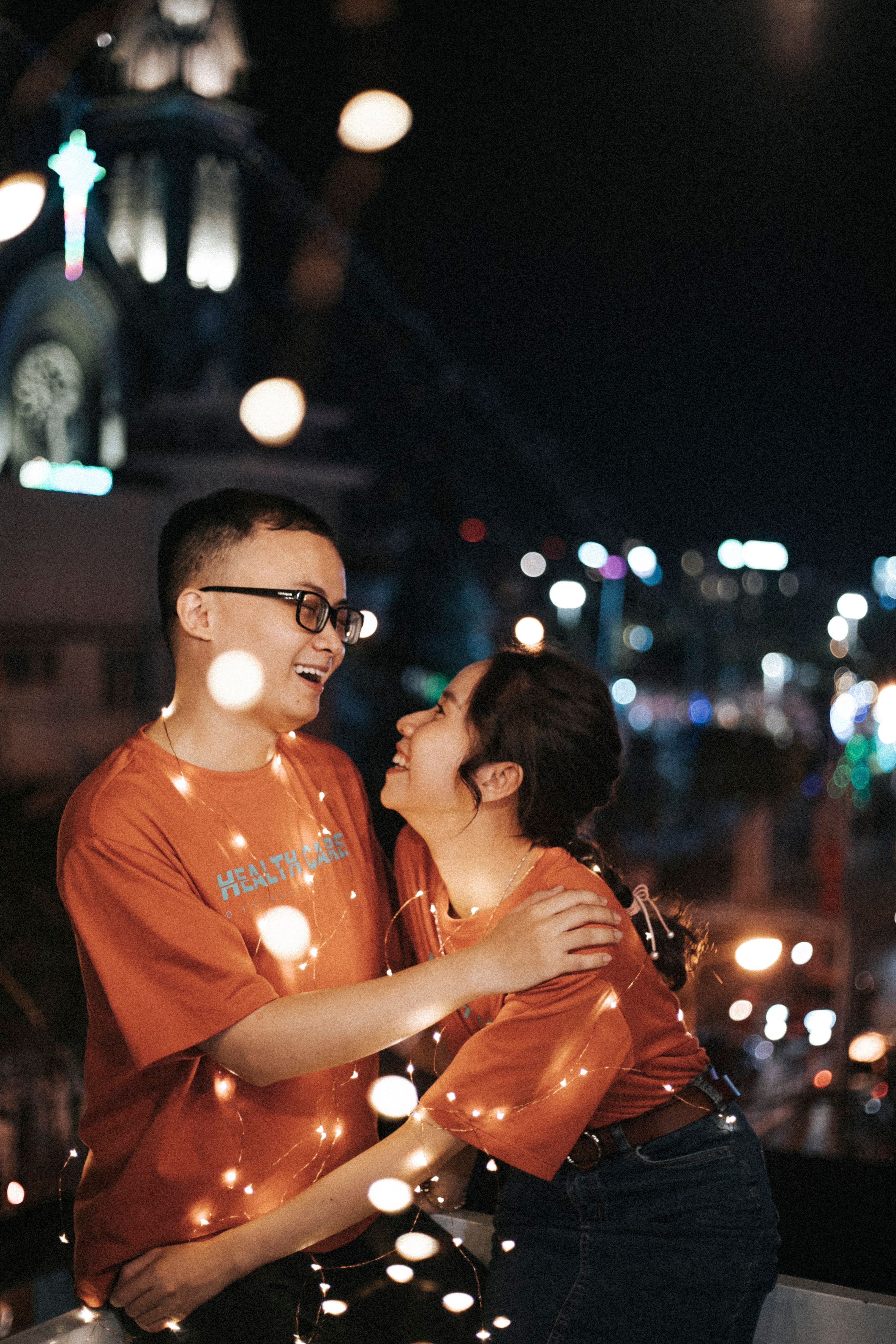 Free Happy couple sharing a joyful moment wrapped in string lights during a night festival. Stock Photo