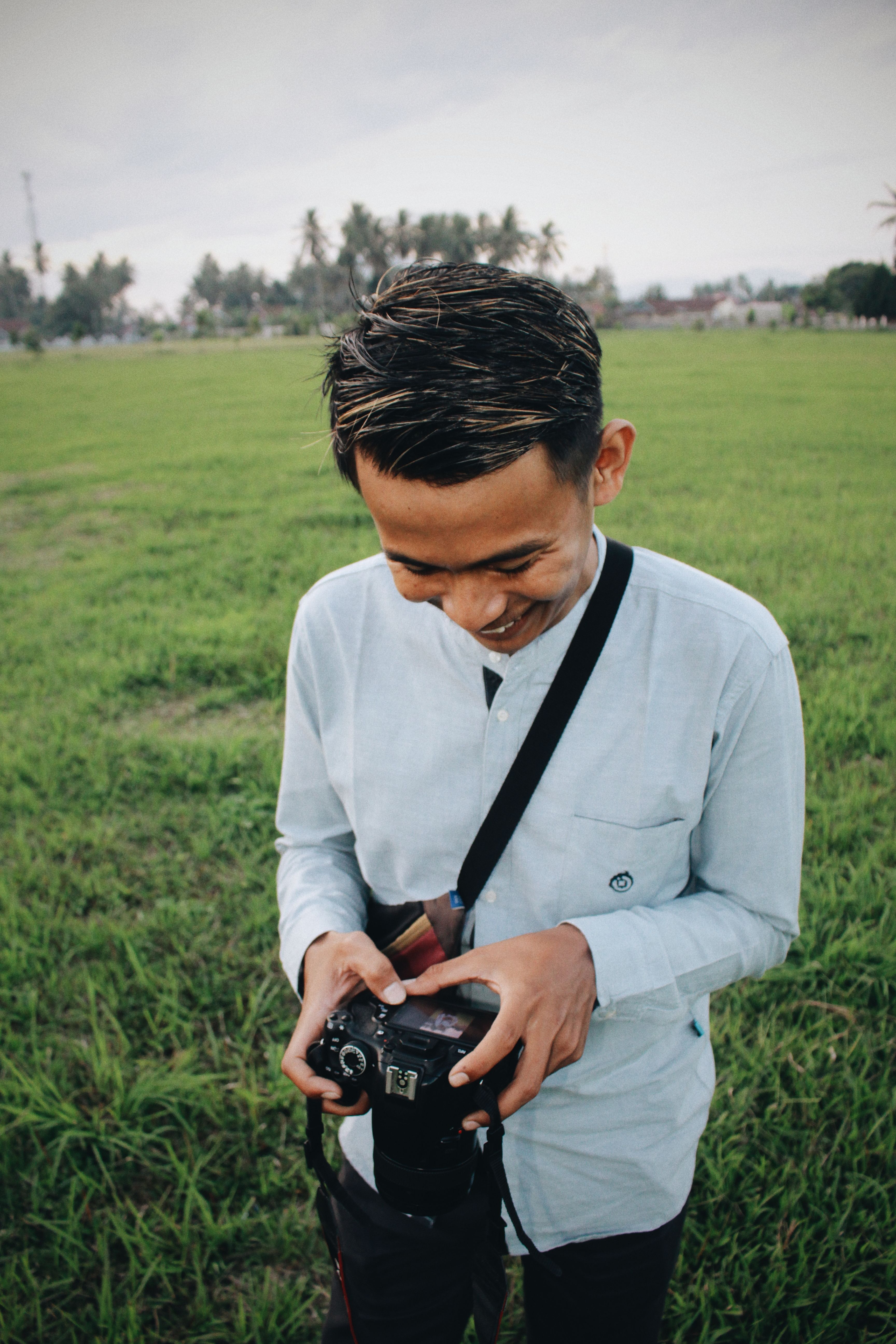 Smiling Asian man checking photos on digital camera while standing on green meadow in tropics