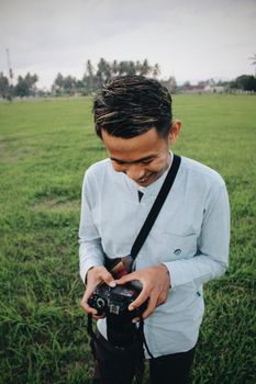 Smiling Asian man checking photos on digital camera while standing on green meadow in tropics