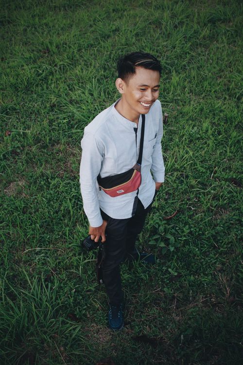 Free Young man with camera bag smiling and standing on grass outdoors. Stock Photo
