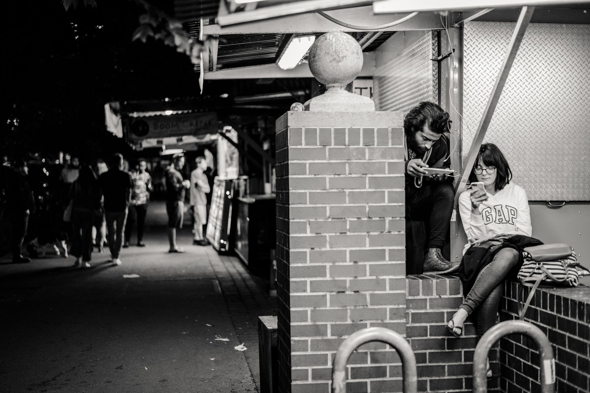 Monochrome photo of people sitting by a brick wall, using smartphones, on a city street.