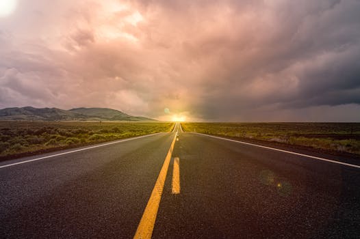 A scenic view of an empty highway stretching towards a sunset horizon under dramatic clouds.