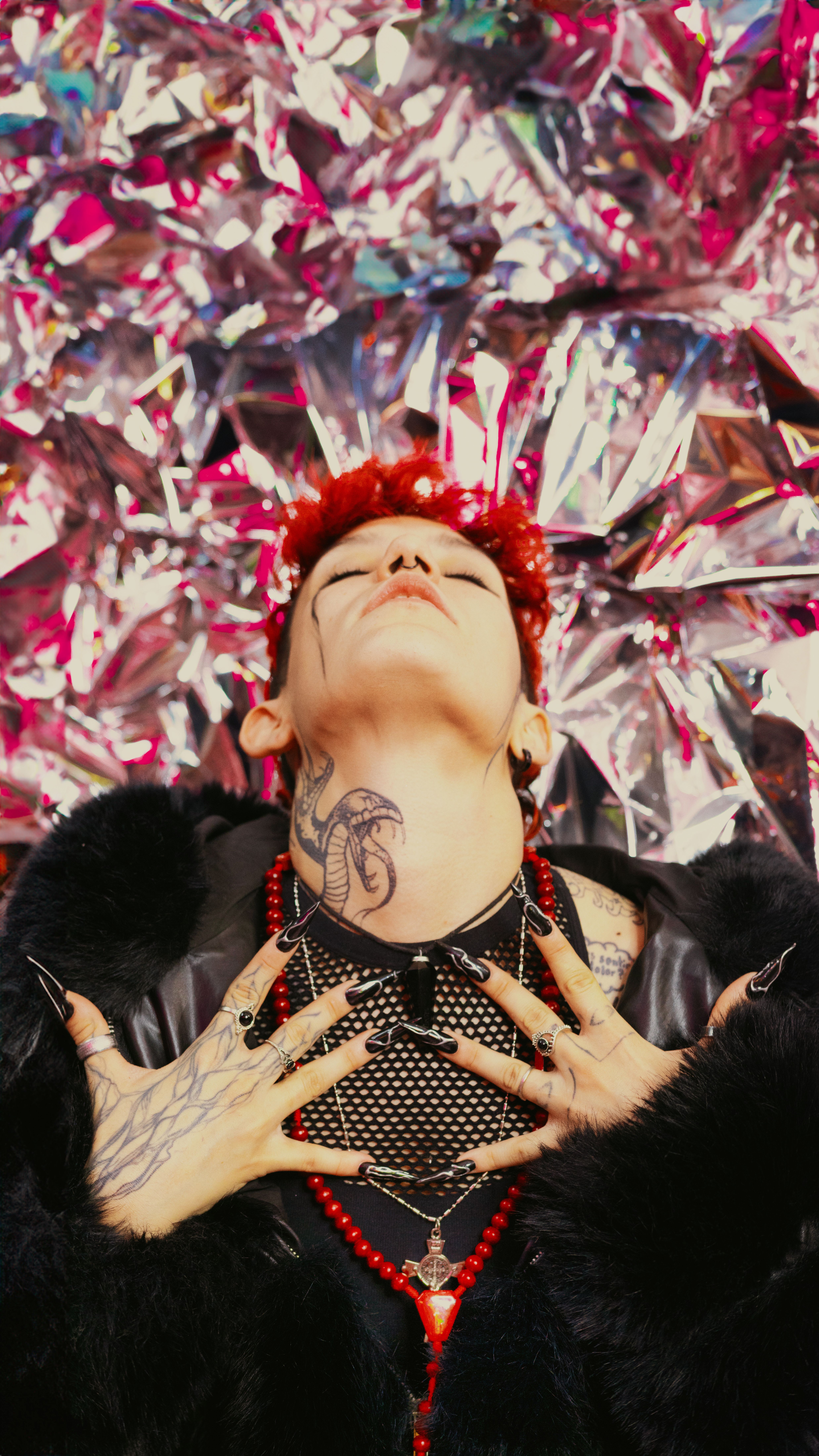 A bold, eccentric individual with striking red hair, tattoos, and a unique manicure poses against a vibrant background.
