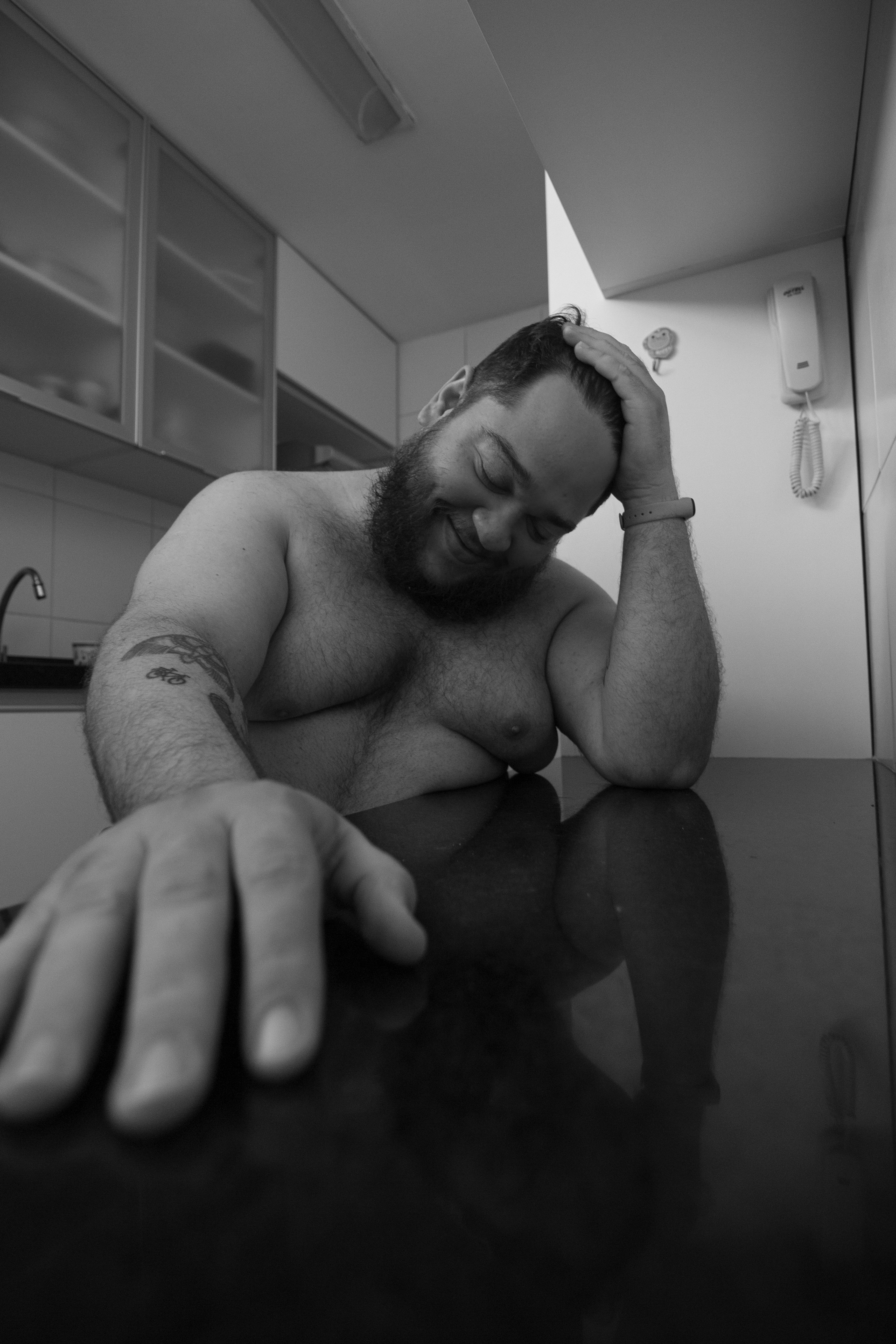 Shirtless man in black and white photo, smiling with hand on table, in a contemporary kitchen setting.