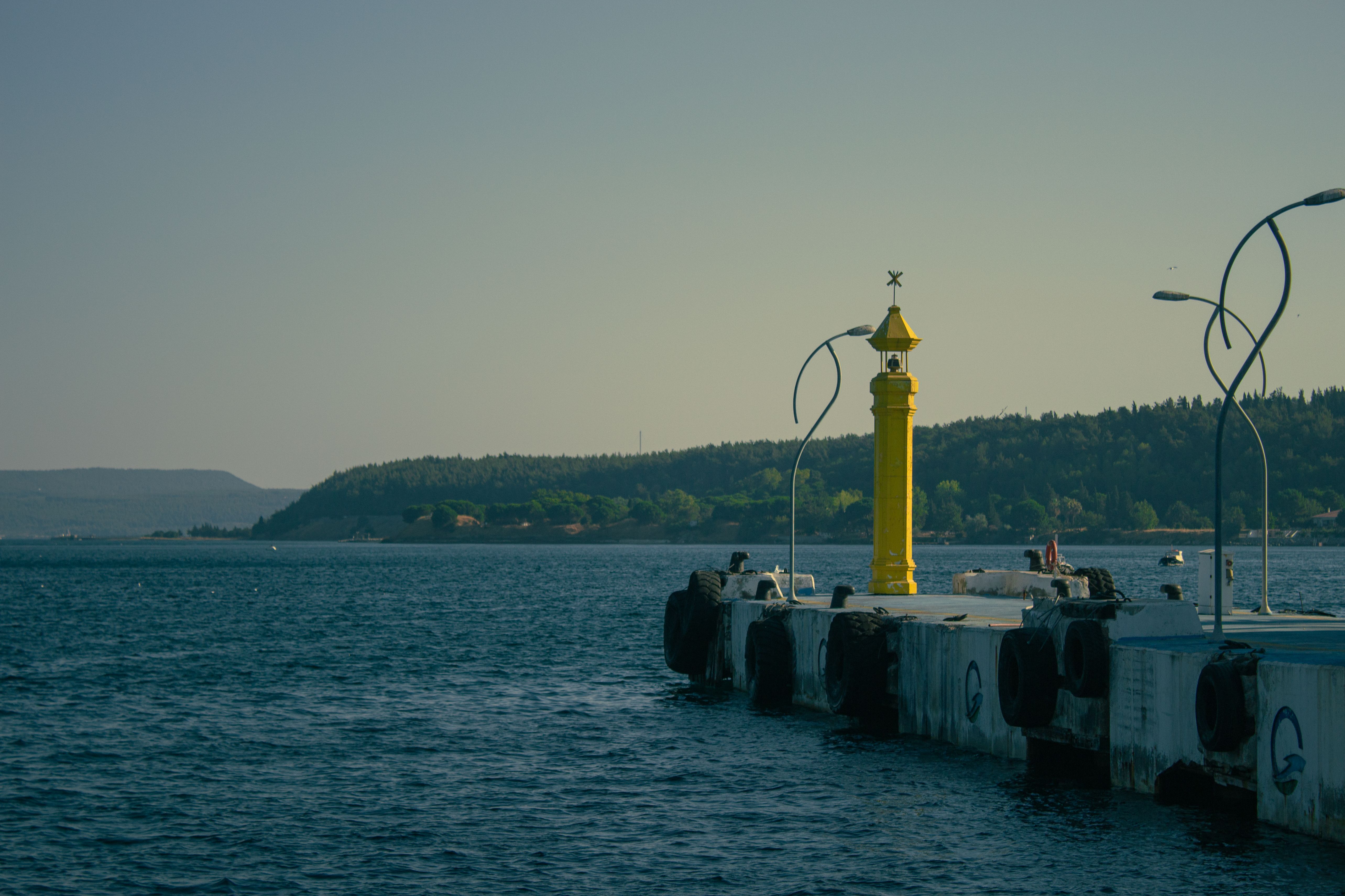 Scenic view of Çanakkale harbor pier with a lighthouse at dusk, capturing serene waters and distant hills.