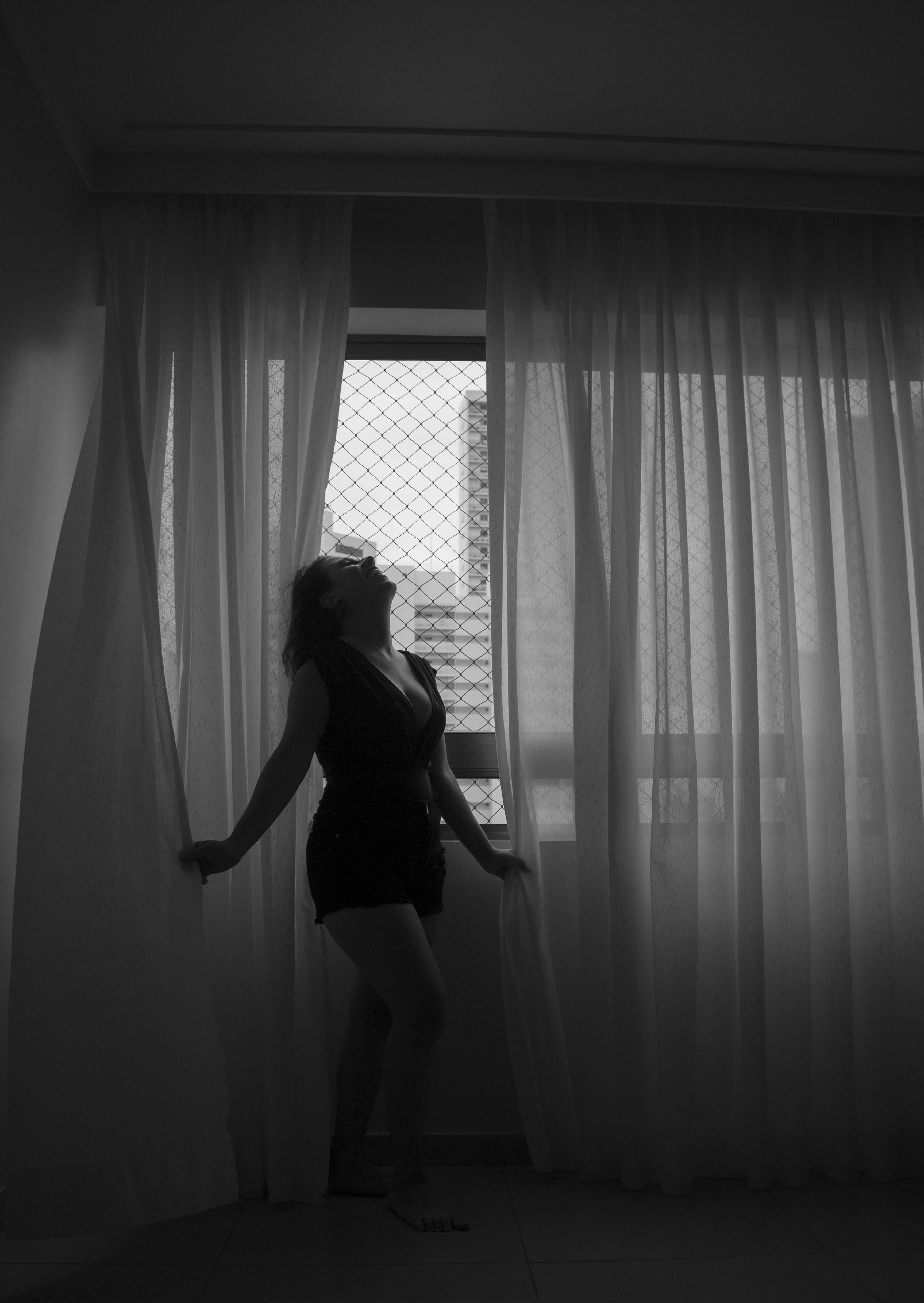 Artistic black and white portrait of a woman silhouetted against window curtains, evoking elegance and mystery.