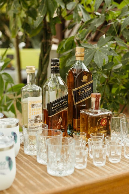 Outdoor setup of assorted liquor bottles and glasses on a wooden table surrounded by greenery.