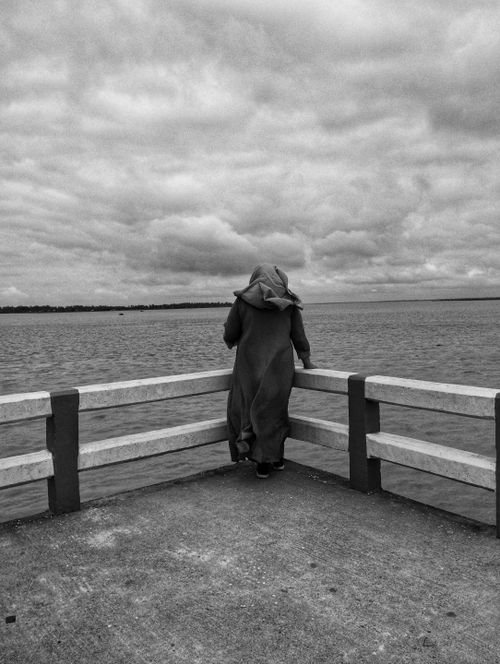 Free Black and white photo of a solitary person standing on a pier, gazing over a stormy sea under cloudy skies. Stock Photo