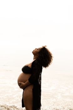 A pregnant woman in a black bikini enjoys a serene moment by the ocean at sunset in São Paulo, Brazil.
