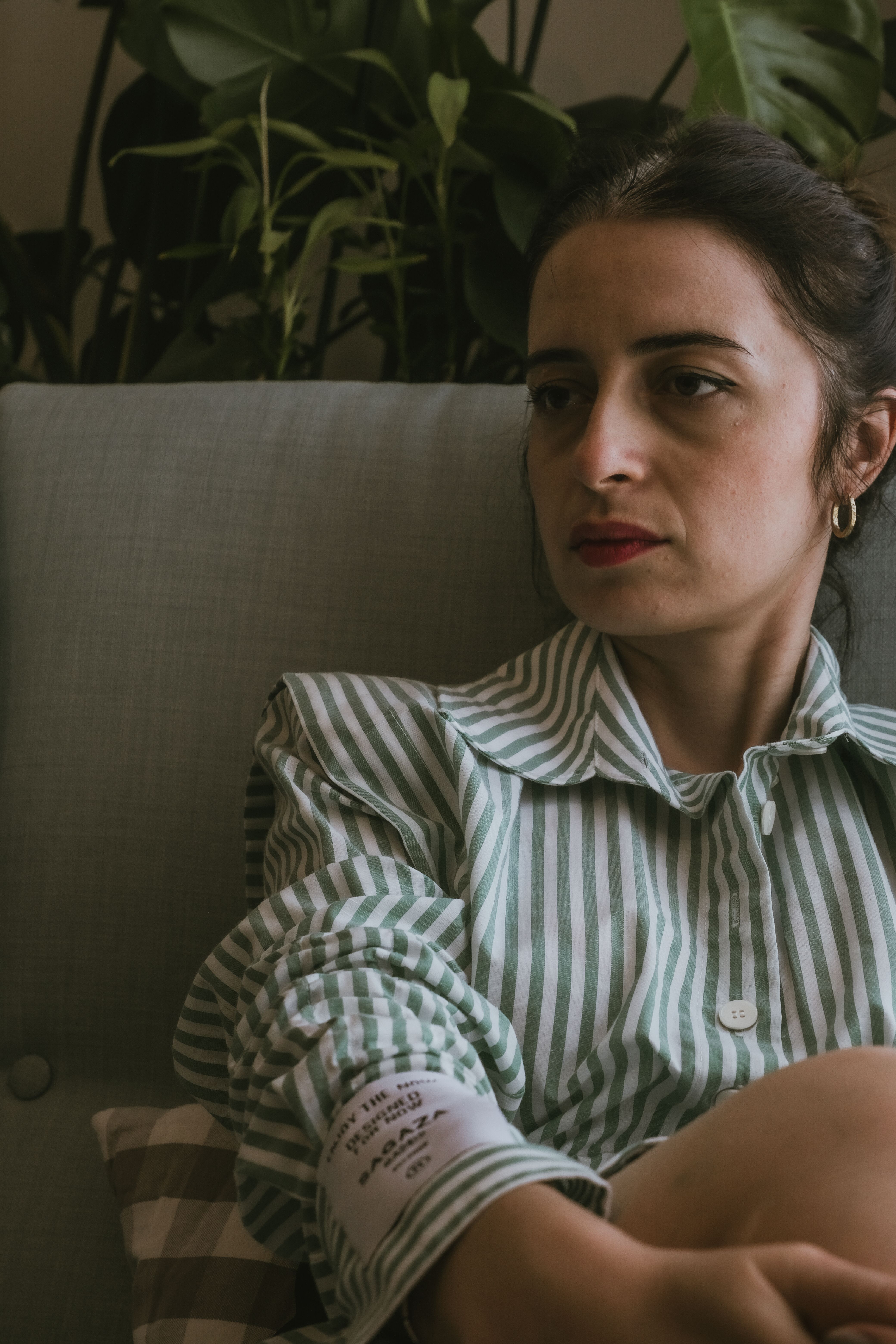 Pensive woman sitting indoors, relaxing on a sofa with plants in the background.