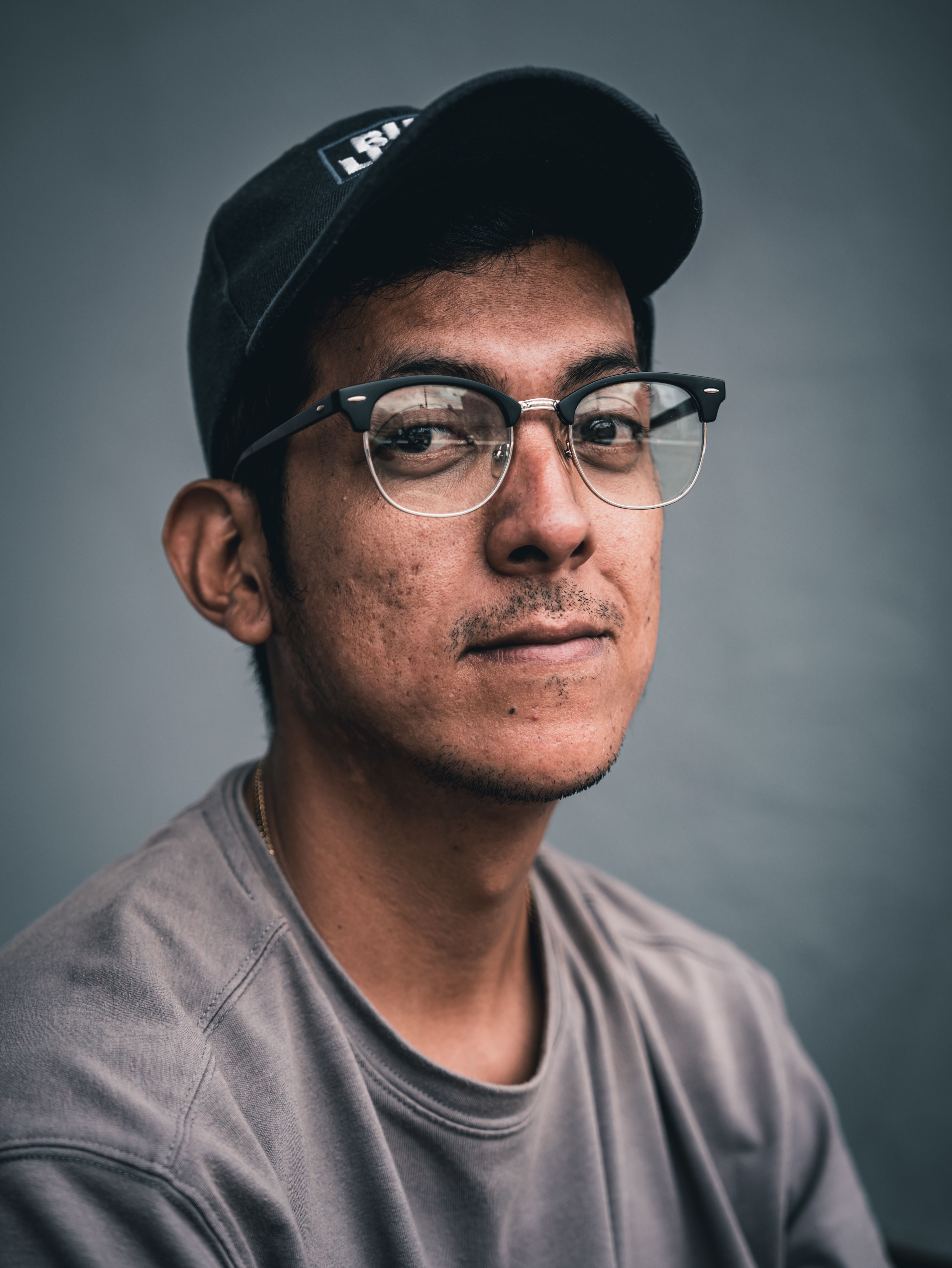 Close-up portrait of a young man wearing eyeglasses and a cap, conveying a serious and pensive mood.