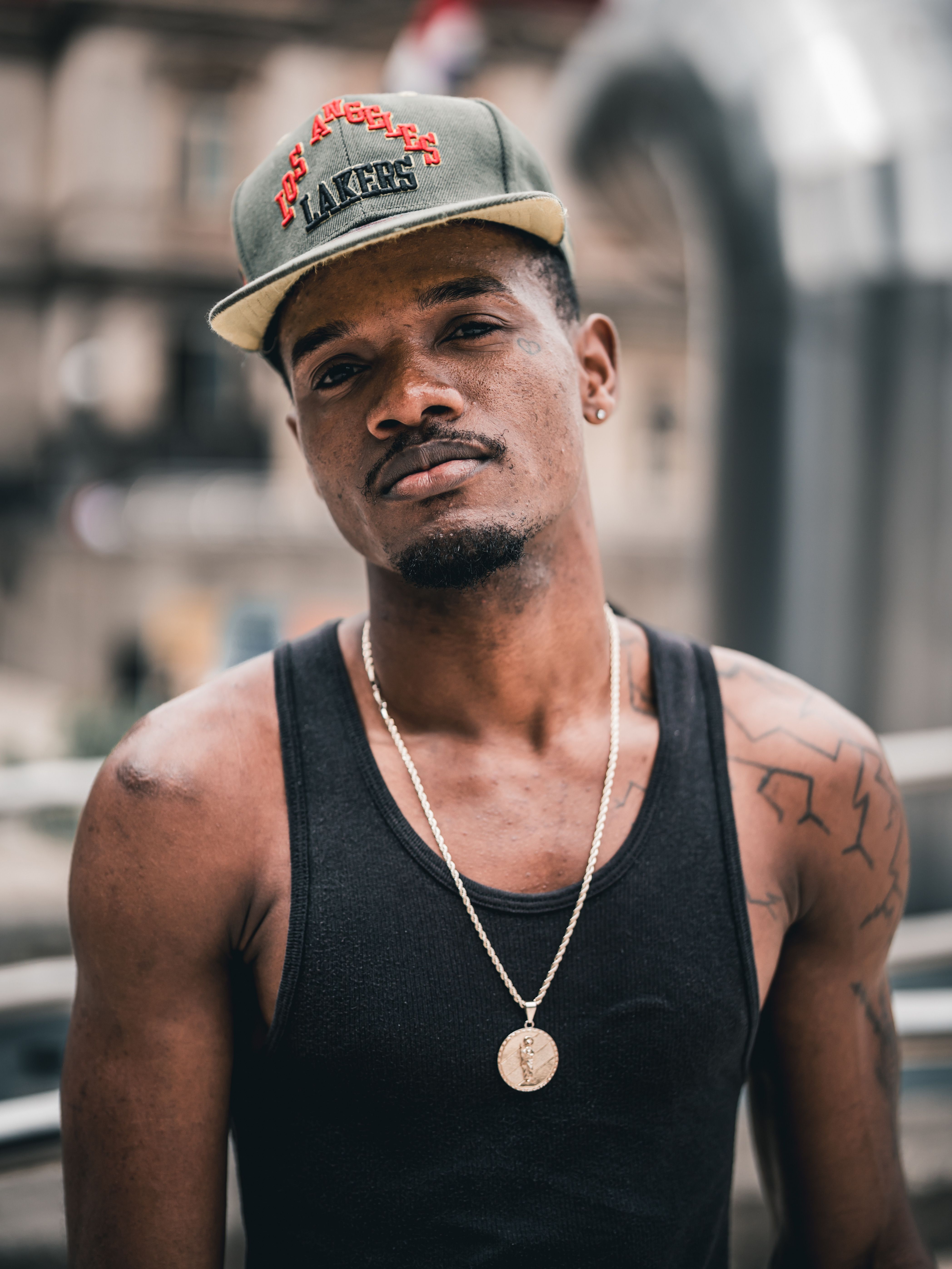 A fashionable adult male wearing a cap and necklace poses confidently outdoors in an urban setting.